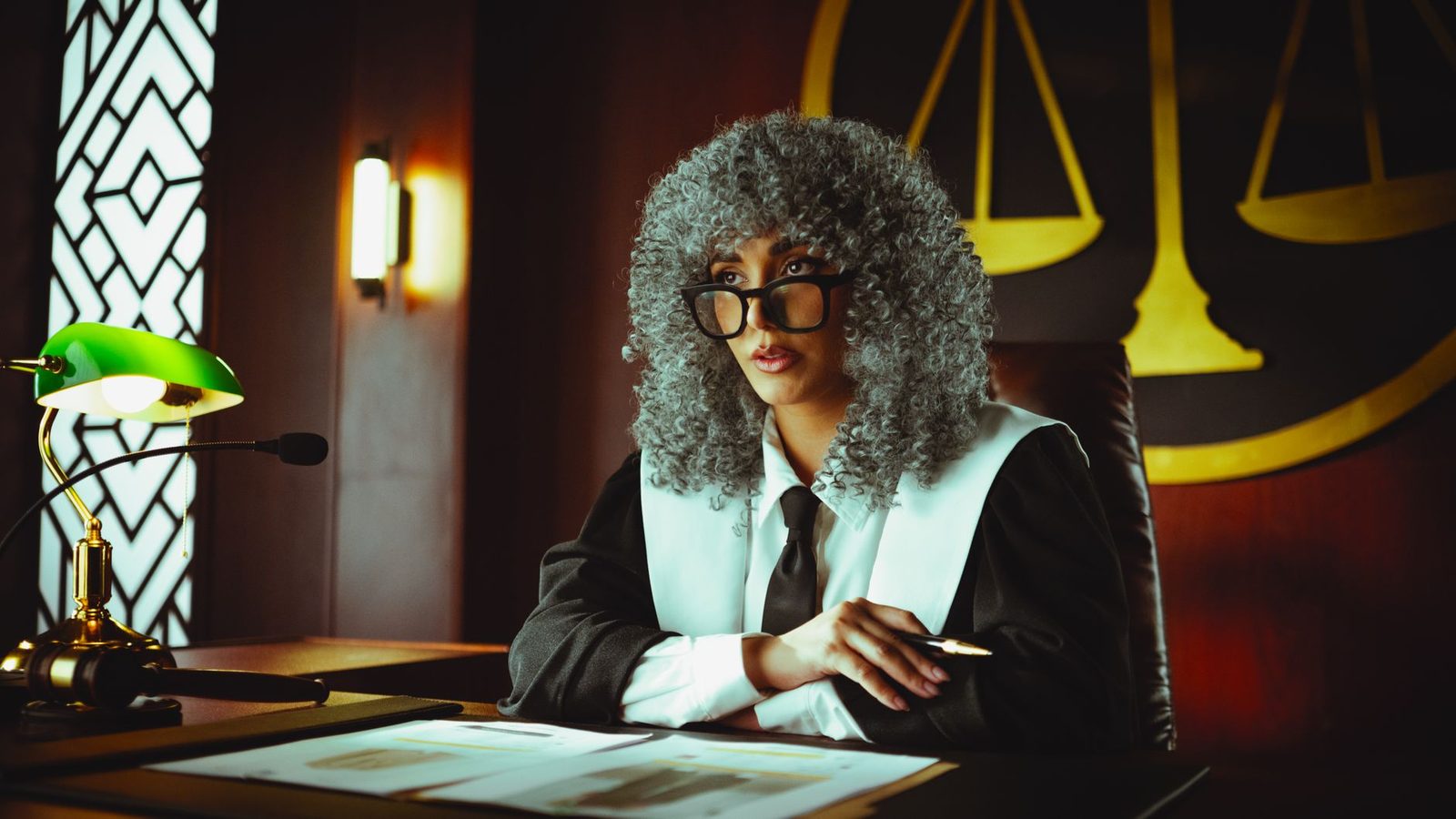 Female judge at courtroom desk with justice scales behind her.