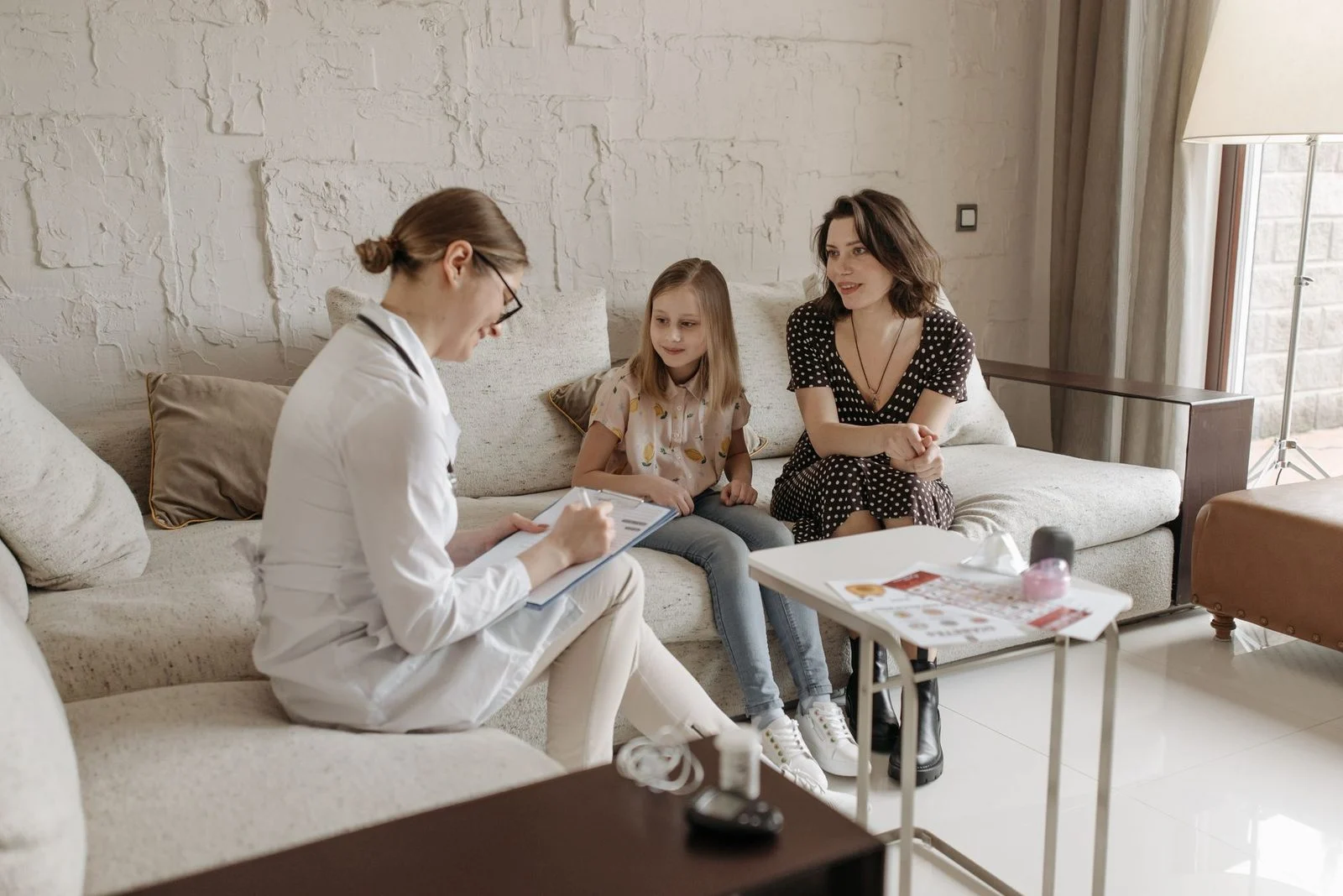 Una médica con bata blanca conversa con una madre y su hija adulta en un sofá acogedor en una sala cálida.
