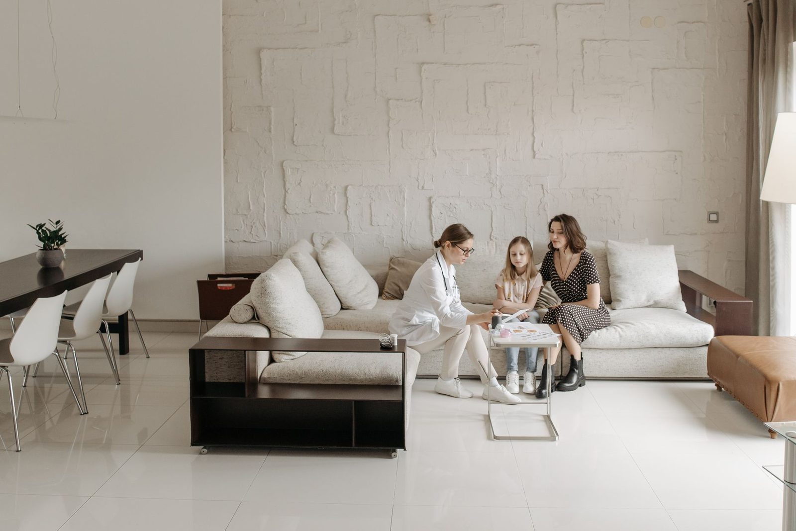A doctor sits with a mother and adult daughter in a warmly lit living room.