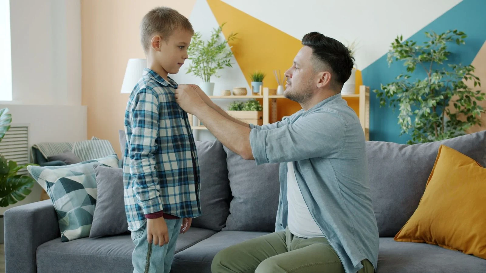 A father helps his adult son fix his shirt collar in a cozy living room.