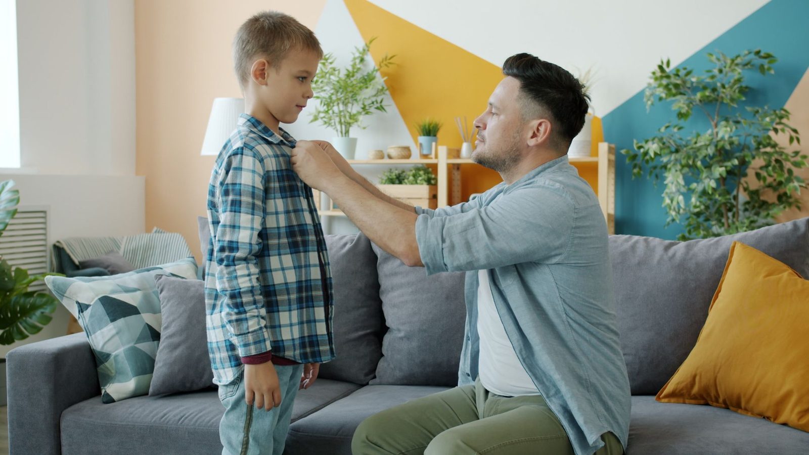 A father straightens his adult son's shirt collar in a cozy living room.