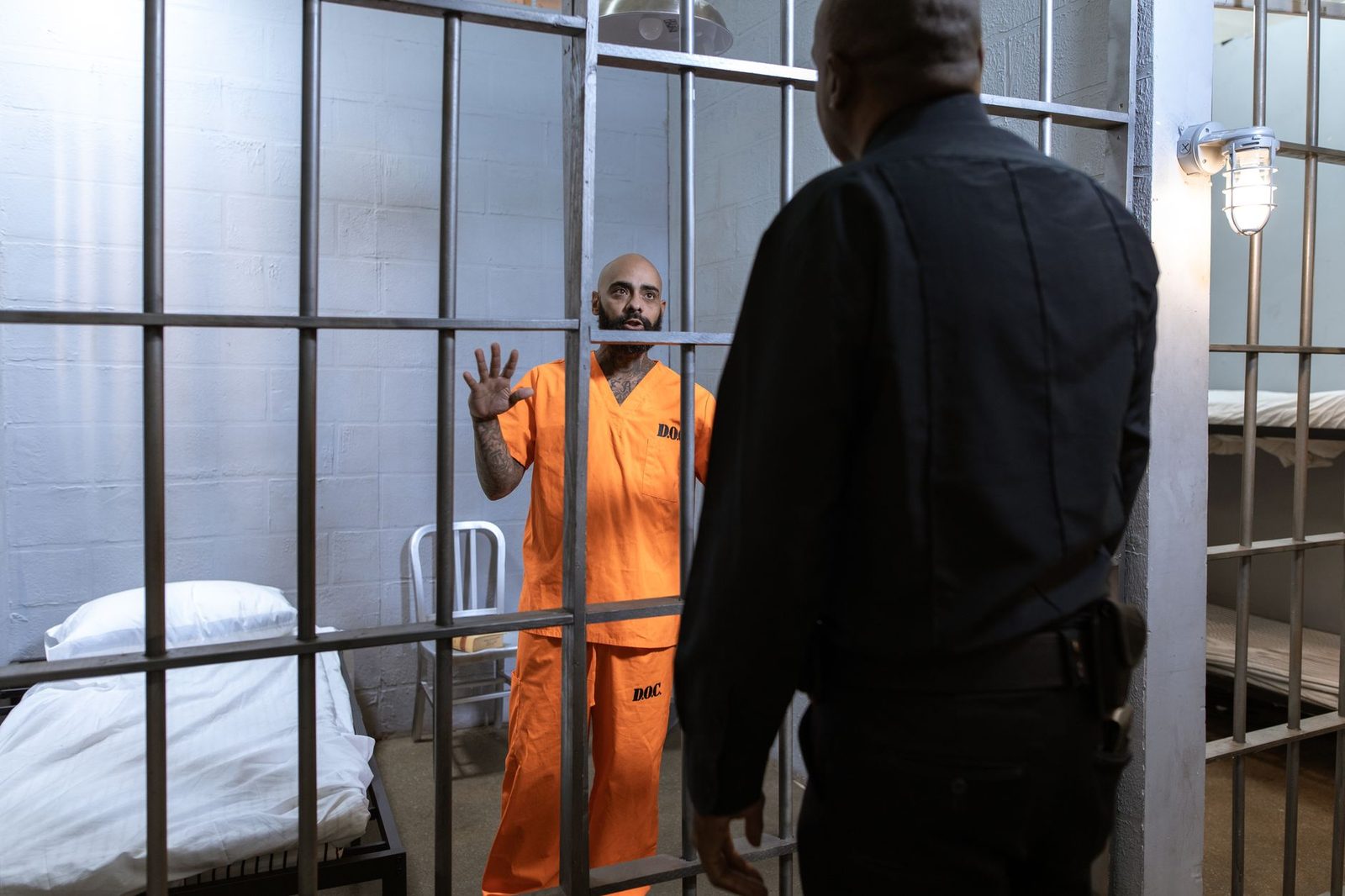 A man in an orange uniform stands behind jail cell bars while a guard looks on.