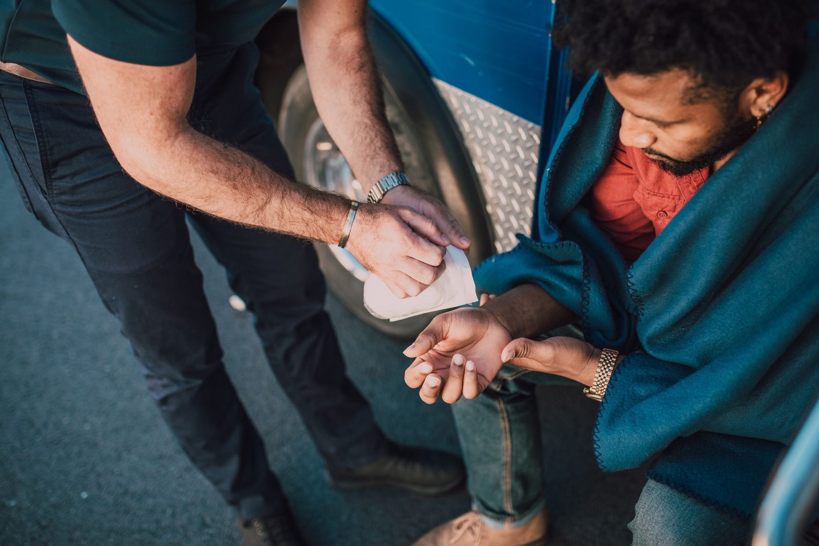 A person kneels beside a man sitting on a city sidewalk, offering first aid to help him.