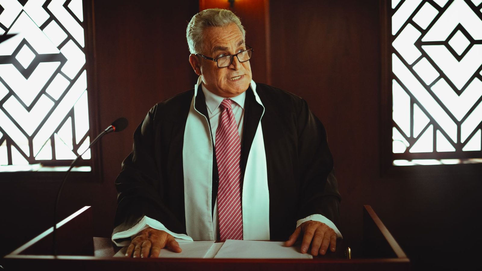 A senior judge in traditional robes stands at the podium in a courtroom.