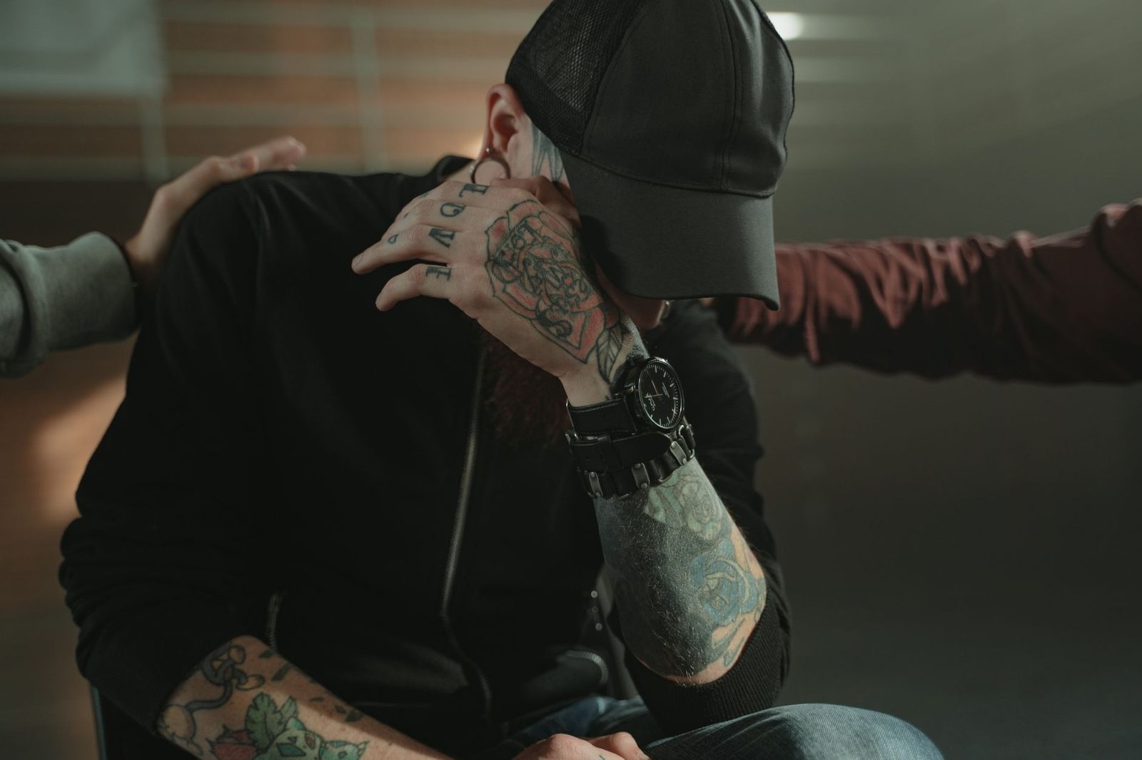 A tattooed person sits indoors while a friend comforts them with a hand on their shoulder.