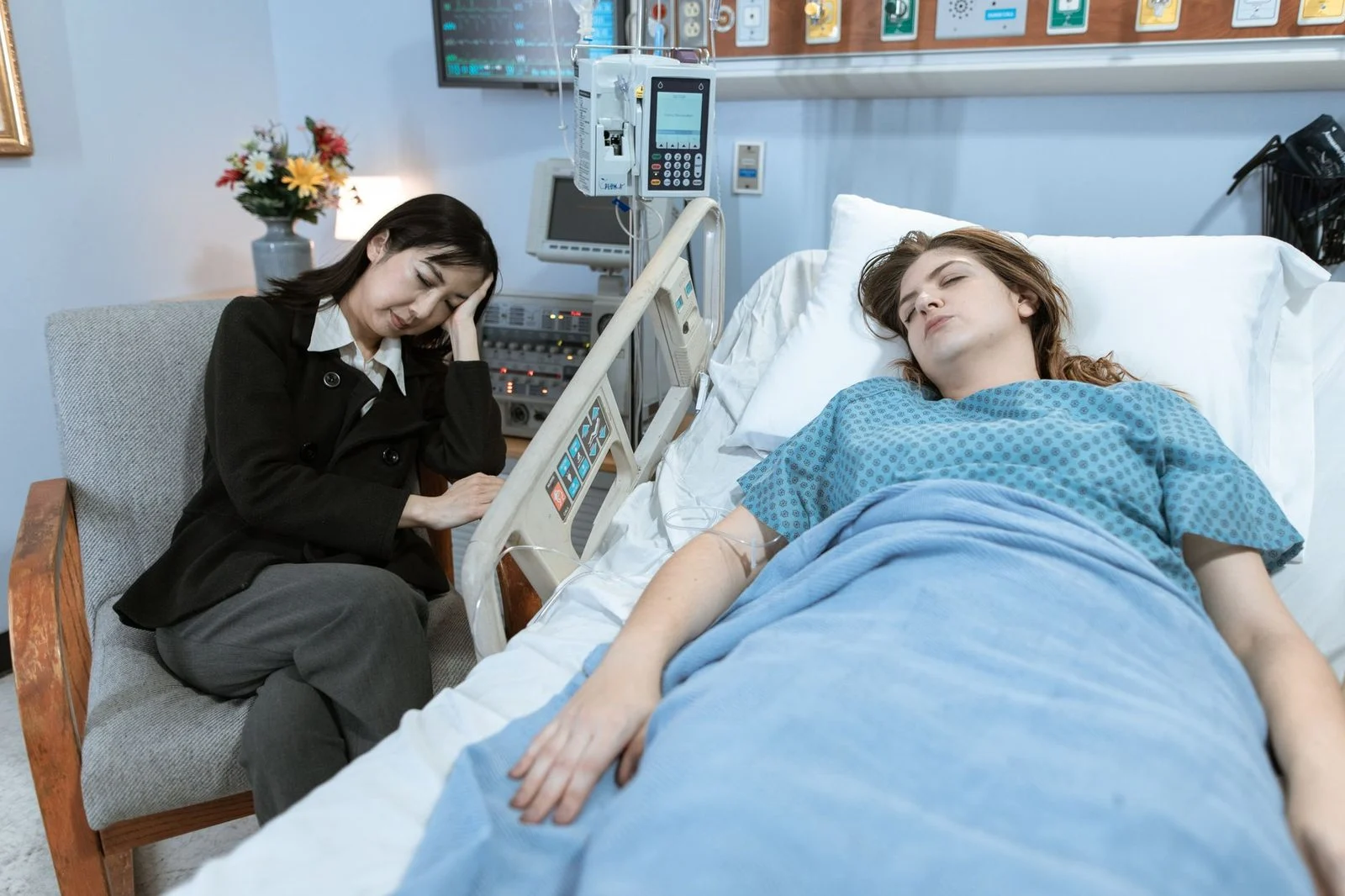 Una mujer preocupada sentada junto a la cama de un hospital mientras un paciente descansa tranquilo.