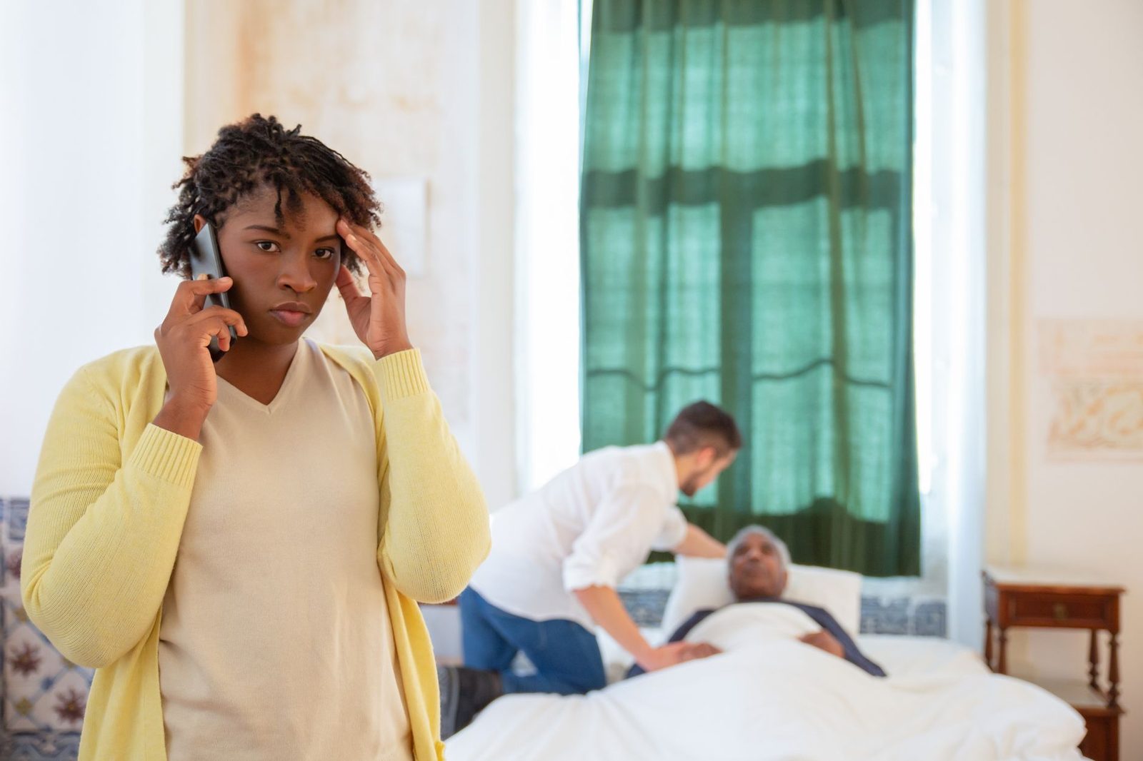 A worried woman talks on the phone while a caregiver helps an elderly person in bed.