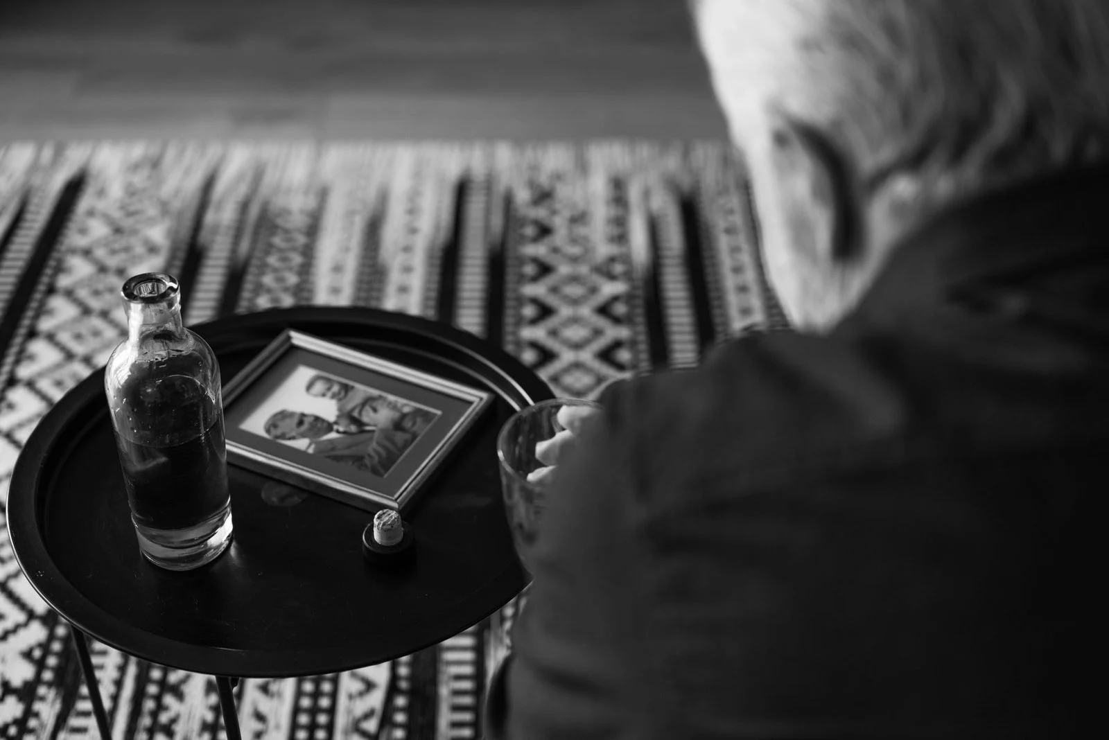 Black-and-white photo of an elderly man sitting thoughtfully beside a framed portrait and a drink.