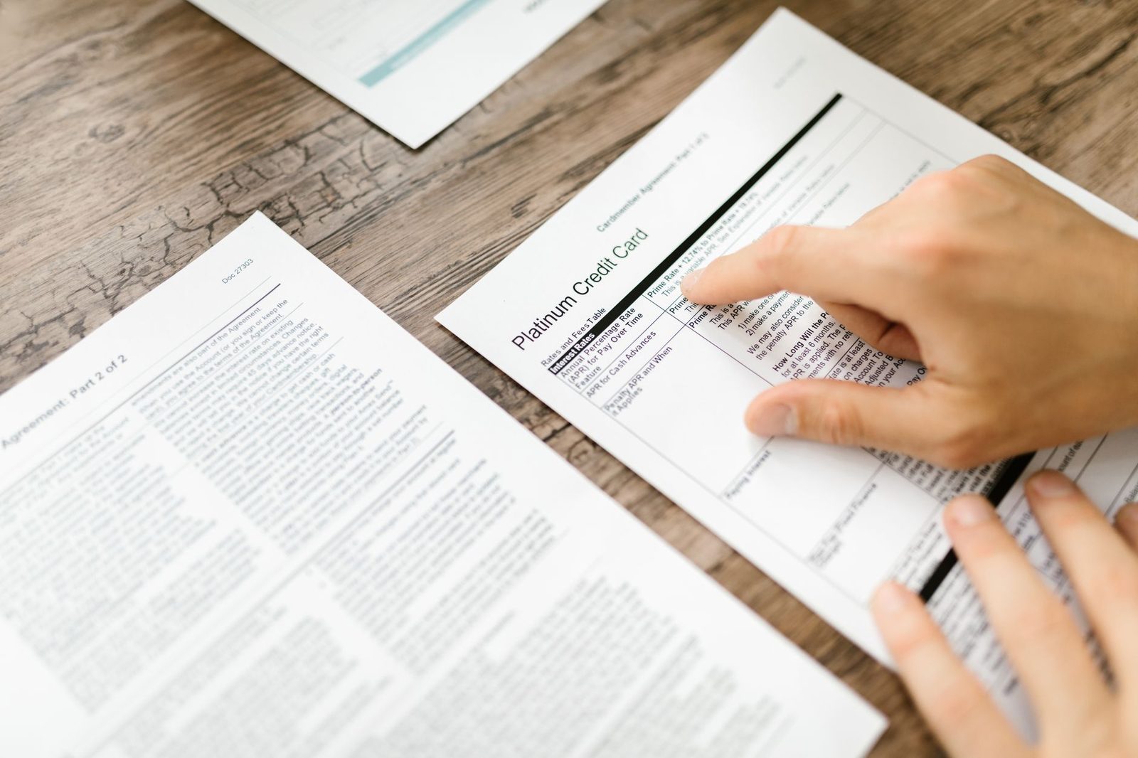 Hand reviewing financial documents and credit card agreement on wooden desk.