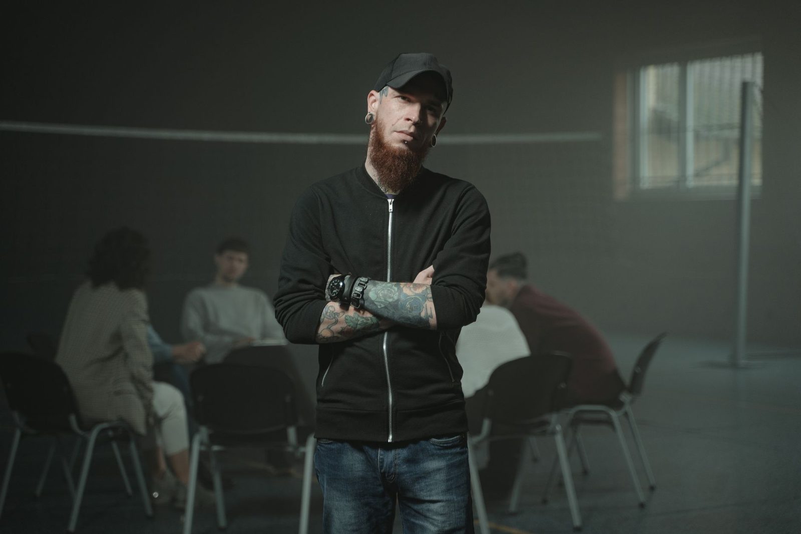 A bearded man with arms crossed stands confidently during a group therapy session.