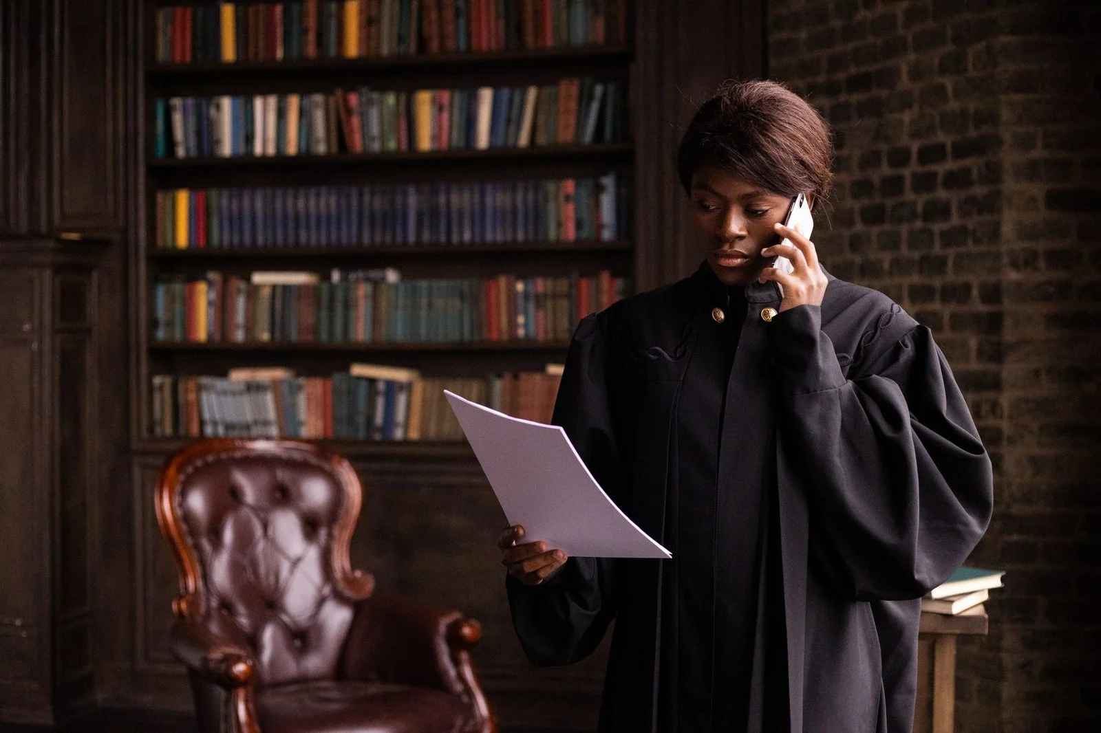 A judge in black robes holds a document and talks on the phone in a law library.