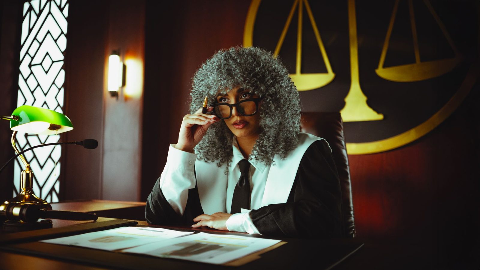 Judge in courtroom at desk wearing traditional robes with serious expression.