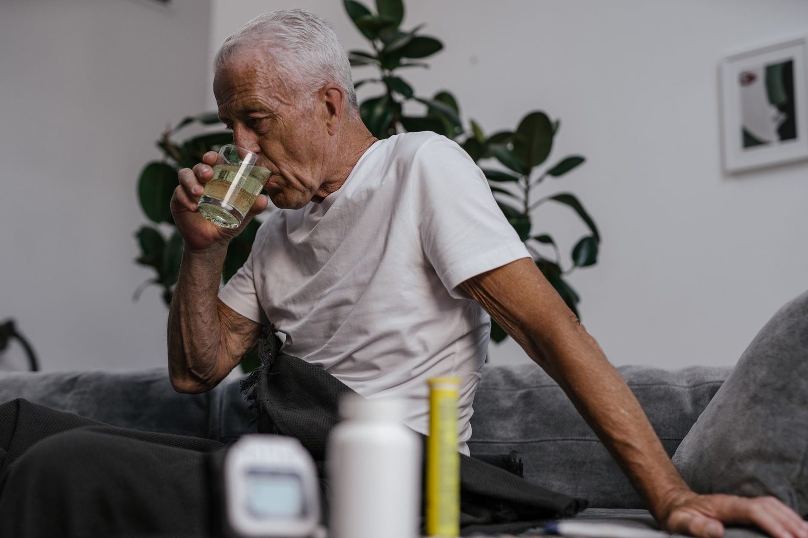 Older man in a white shirt takes a drink indoors, possibly medicine.