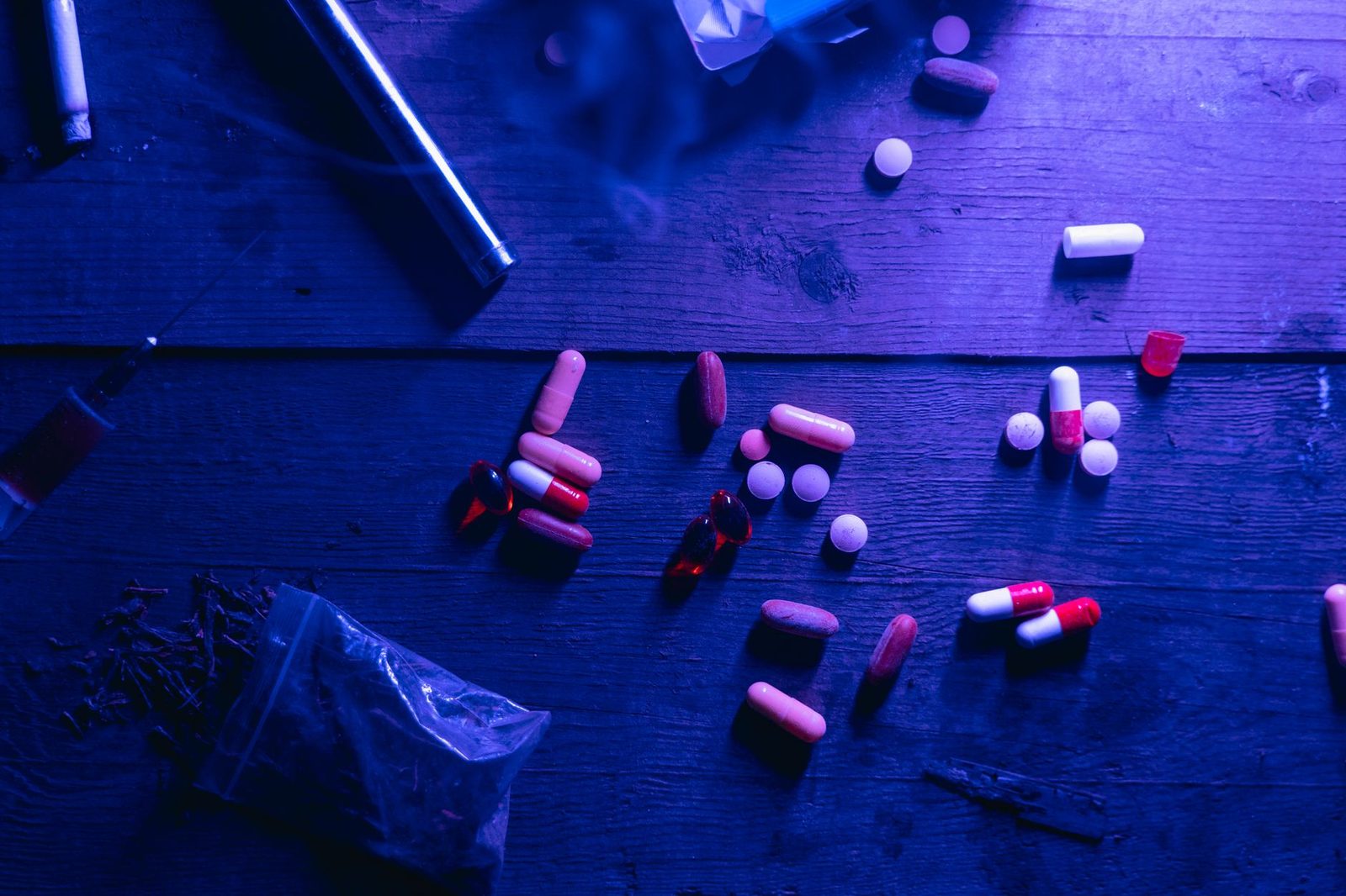 Pills, capsules, and a syringe arranged on a wooden table under blue lighting.