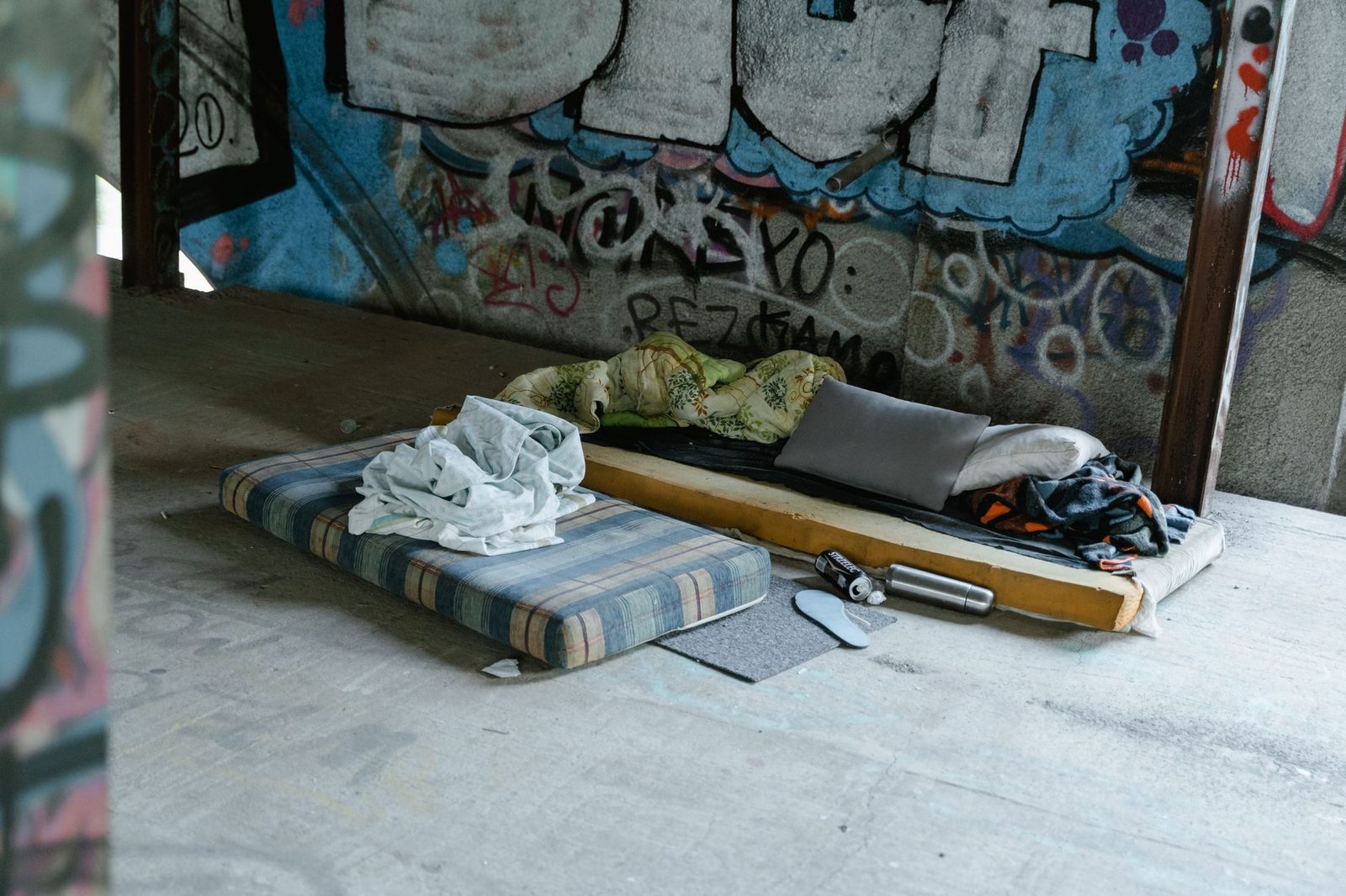Refugio urbano abandonado con colchón, grafiti y vaso metálico en el suelo.
