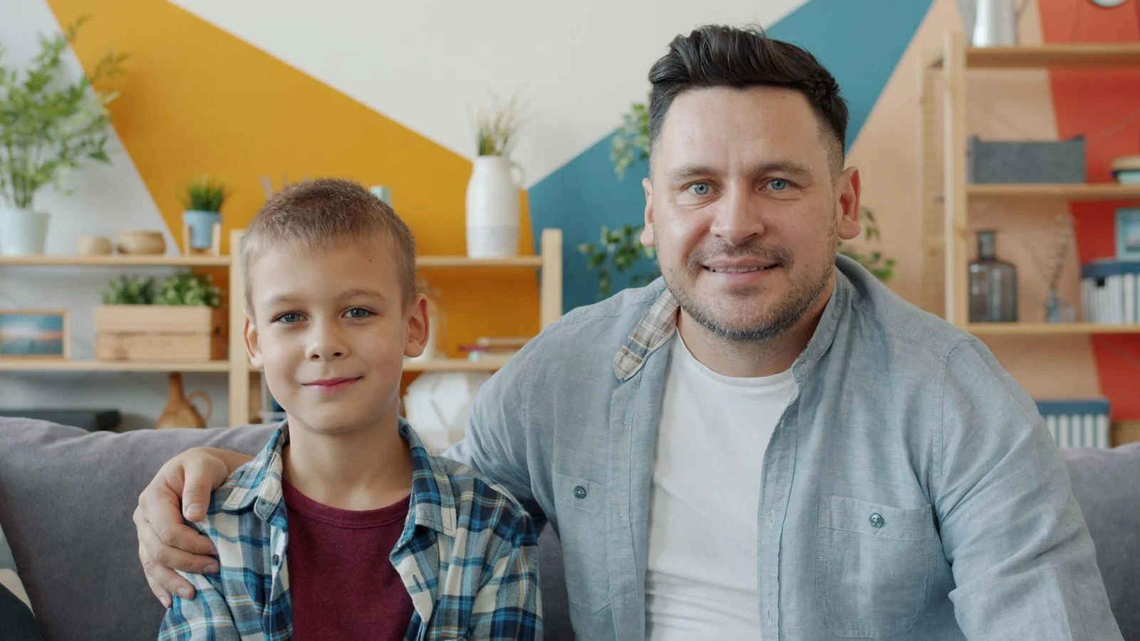 Padre sonriente sentado junto a su hijo adulto en una sala con decoración moderna y colorida.