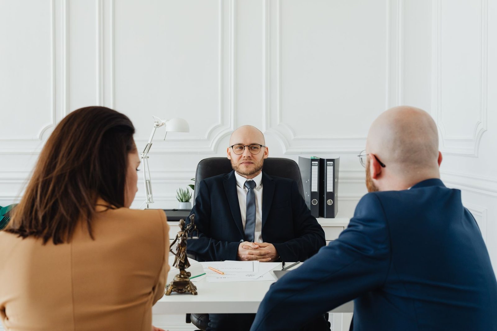 Two adults sit across a desk from a lawyer in a formal office, reviewing papers during a legal consultation.