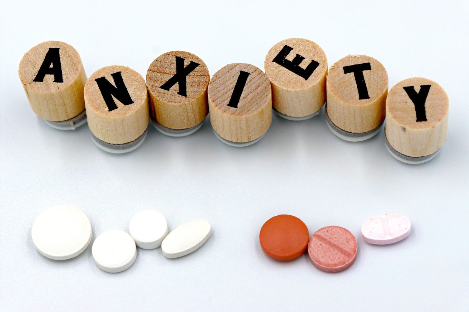 Bloques de madera que deletrean "Anxiety" sobre pastillas esparcidas en un fondo claro.