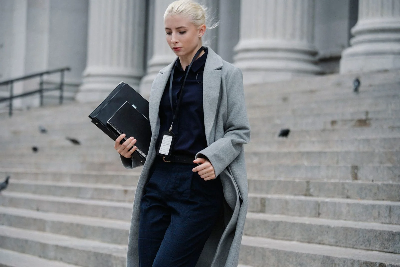 Young woman in formal attire holding folders while walking down stone steps past a columned building.