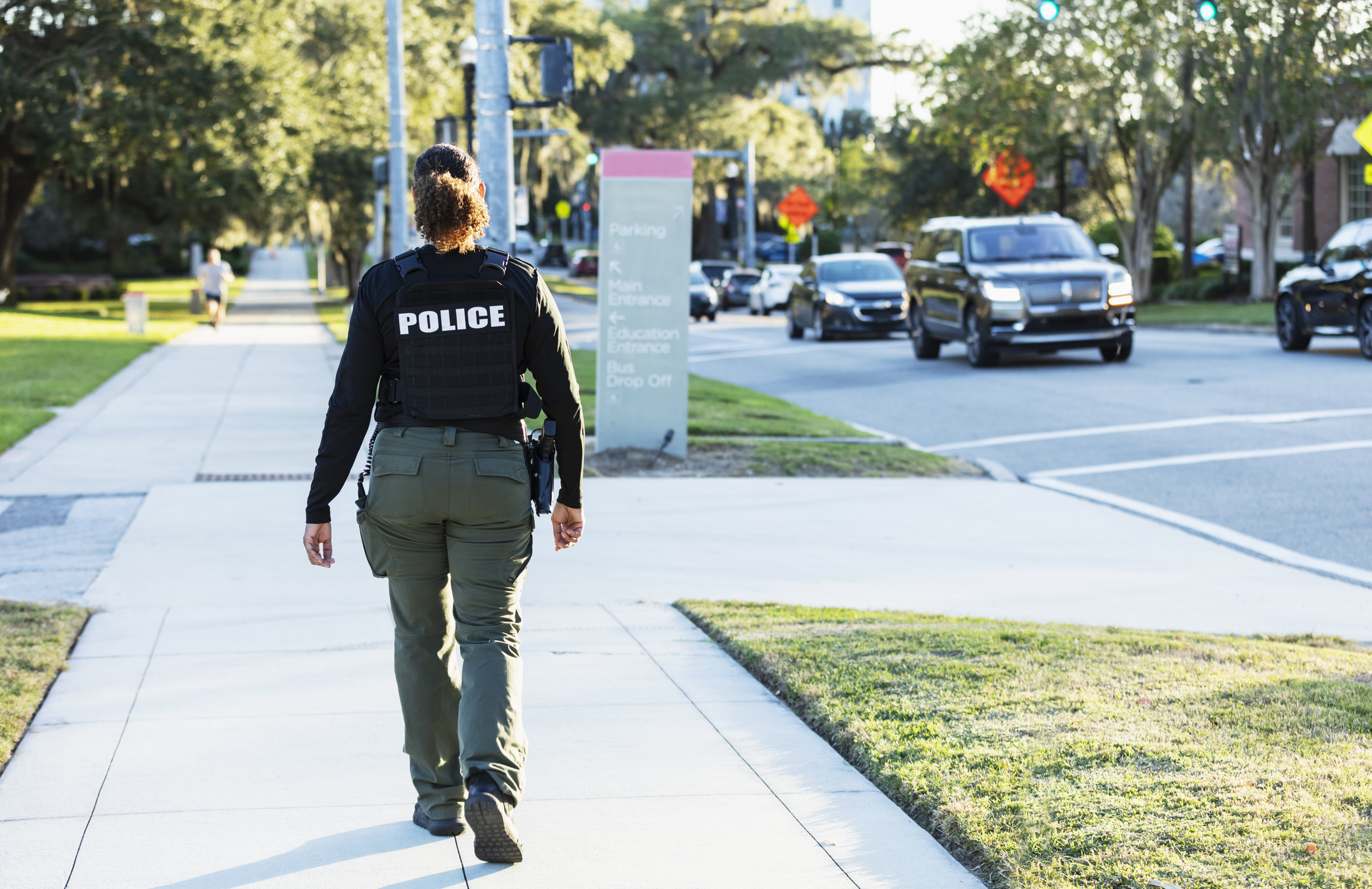 Rear view of a mature multiracial policewoman patrolling an urban area on foot. She is walking on a sidewalk away from the camera.